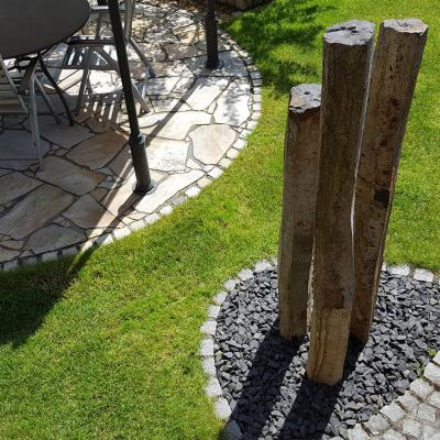 Drei Basalt-Steinsäulen stehen in einem kleinen Kiesbeet neben einer Ruhezone im Garten. Diese dienen als Blickfang in einem gepflegten Garten.