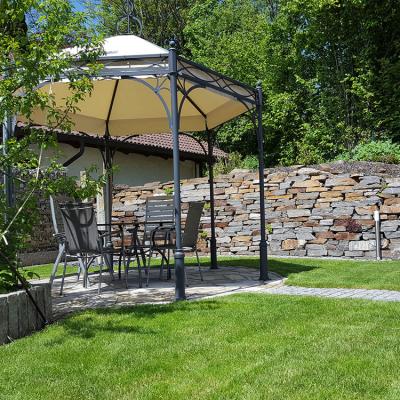 Ein Pavillon mittig auf einer Gartenanlage. Umgeben von einer gepflegten Rasenfläche. Dahinter eine große Natursteinmauer.