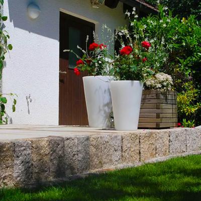 Eine Terrasse mit sauberen Randabschluss durch Naturstein. Auf der Terrasse befinden sich zwei weiße Tontöpfe mit Rosen.
