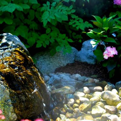 Eine künstlich angelegte Quelle im Steingarten. Aus einem weißbraunen Stein quillt Wasser auf eine Kiesfläche.