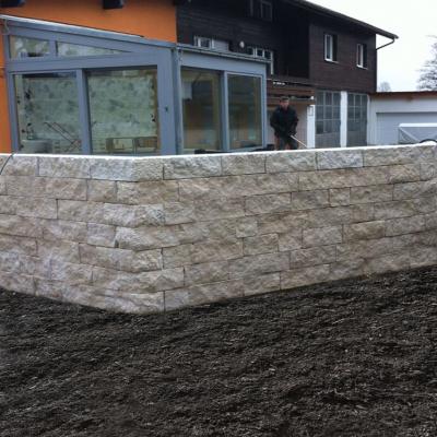 Eine helle rechtwinkelige siebenreihige Mauer aus Blocksteinen vor einer Terrasse.