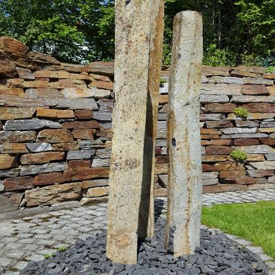 Drei Steinsäulen in einem kleinen Kiesbeet zur Verzierung des Gartens. Dahinter eine hohe Mauer mit Natursteinplatten.