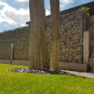 Eine grüne Rasenfläche mit drei Steinsäulen in einem kleinen Kiesbeet. Dahinter steht eine lange hohe Steinmauer (Gabione).